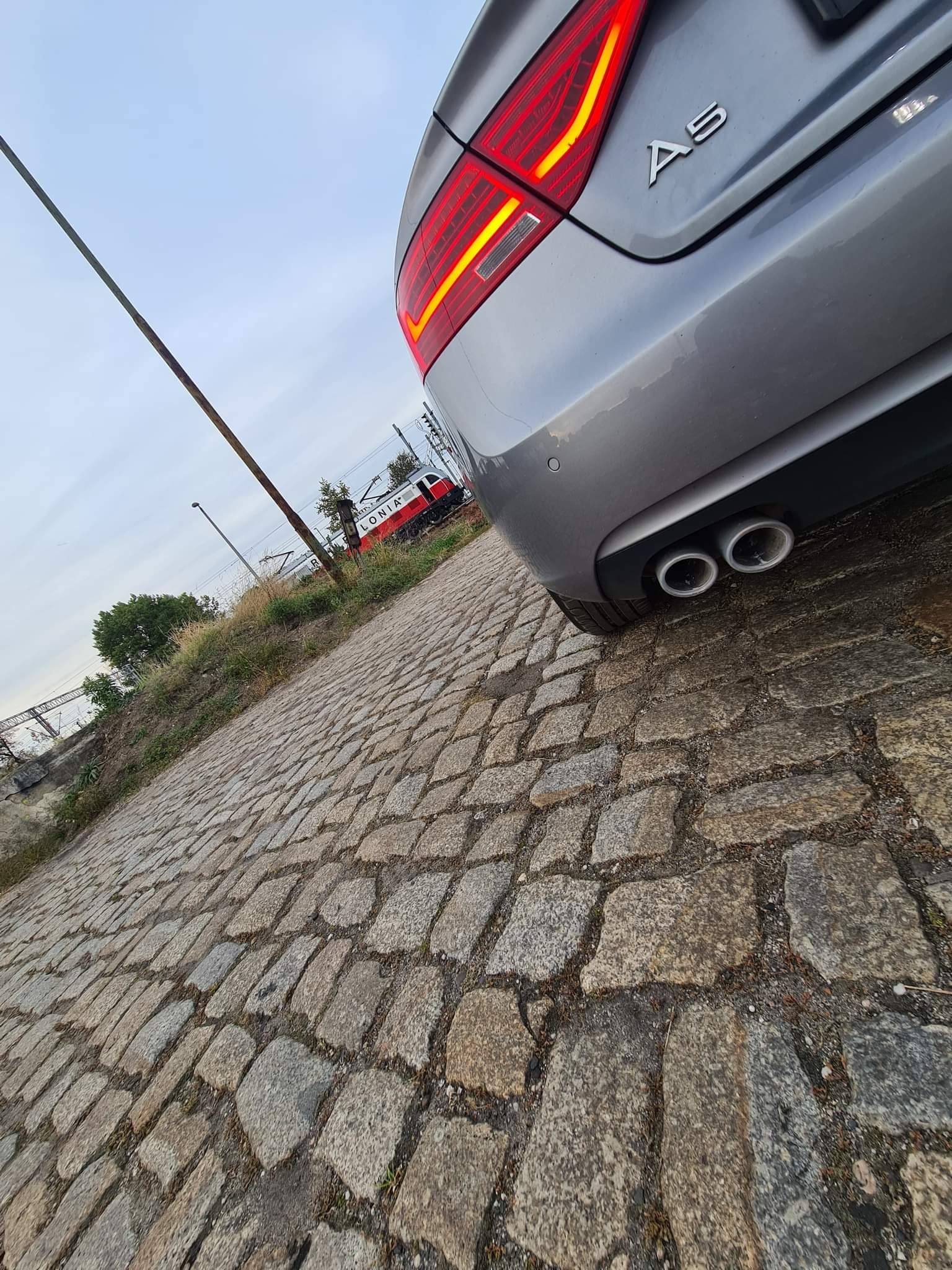 Tył srebrnego samochodu Audi A5 z widocznymi światłami i podwójnym wydechem, zaparkowanego na brukowanej drodze w Radwanicach, z pociągiem w tle.