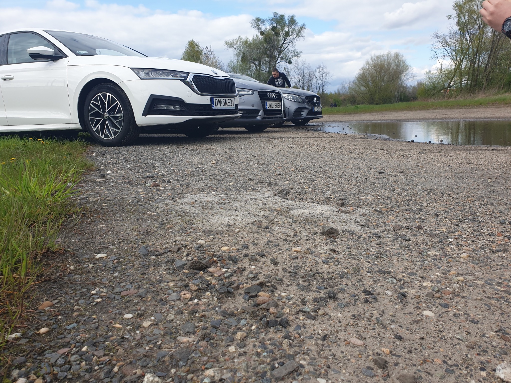 Trzy samochody (Skoda, Audi, Mercedes) zaparkowane na żwirowym terenie z kałużą w tle, osoba w czarnej bluzie przy Audi.