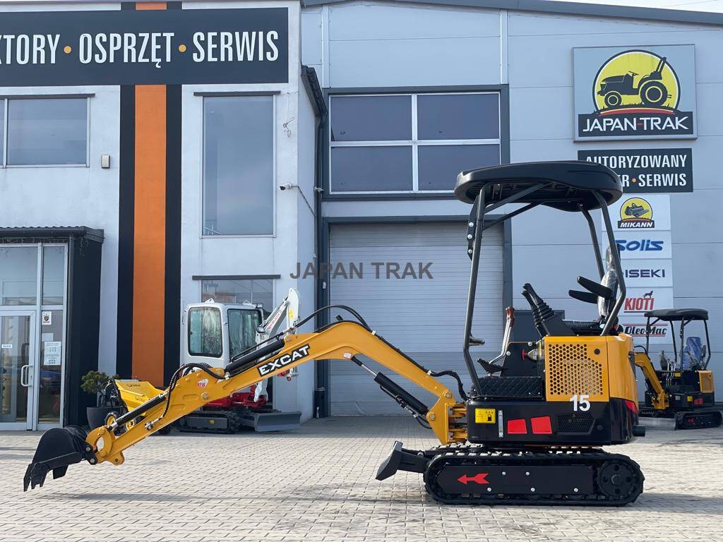 Żółto-czarna maszyna budowlana EXCAT z łyżką z przodu, stojąca na placu przed budynkiem z szyldem JAPAN TRAK, w tle widoczne inne maszyny.