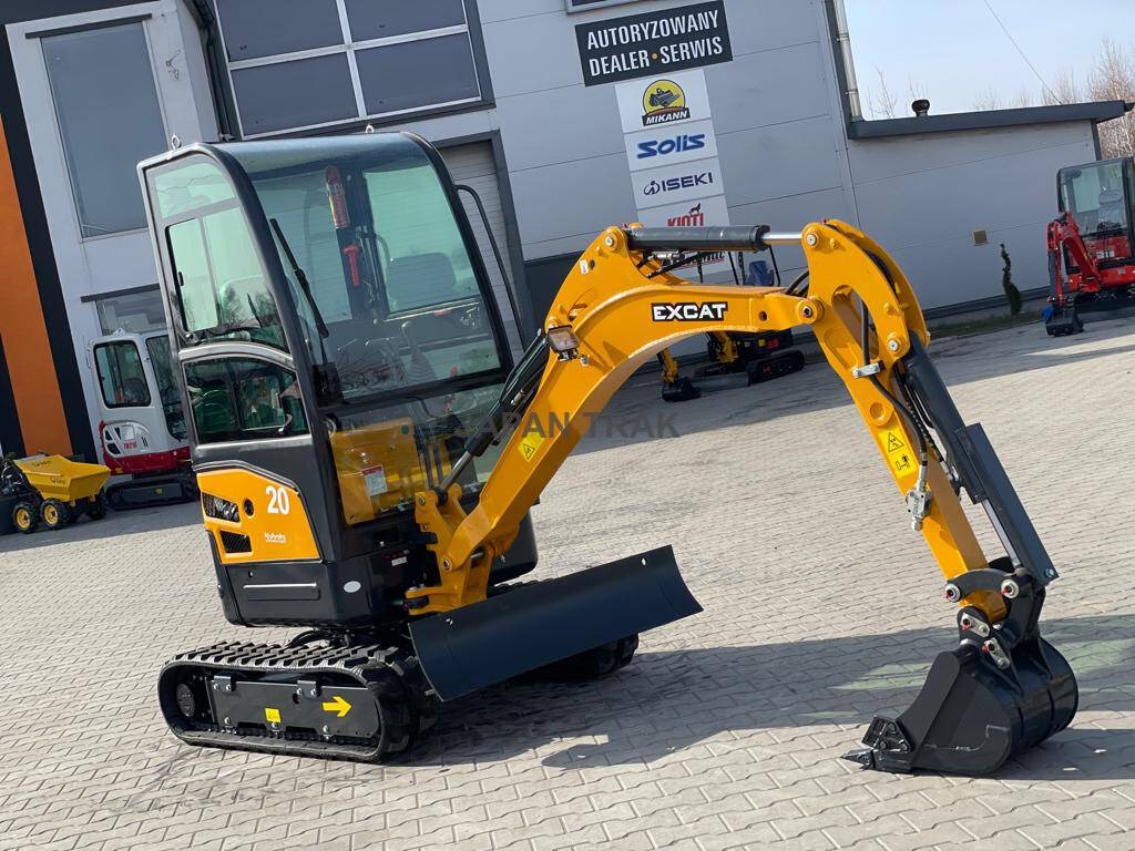 Żółto-czarna, fabrycznie nowa, mała koparka EXCAT 20 na gąsienicach, stojąca na kostce brukowej przed budynkiem z logo autoryzowanego dealera i serwisu.