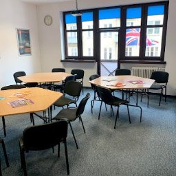 The London School of English - Sala lekcyjna z trapezoidalnymi stolikami, czarnymi krzesłami, rzutnikiem pod sufitem i widokiem na budynek z flagą brytyjską za oknem.