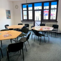 Sala lekcyjna z trapezoidalnymi stolikami, czarnymi krzesłami, rzutnikiem pod sufitem i widokiem na budynek z flagą brytyjską za oknem.