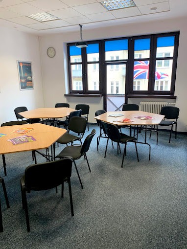 Sala lekcyjna z trapezoidalnymi stolikami, czarnymi krzesłami, rzutnikiem pod sufitem i widokiem na budynek z flagą brytyjską za oknem.