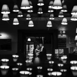 Czarno-białe zdjęcie pary młodej obejmującej się pod dekoracyjnym żyrandolem w eleganckim wnętrzu hotelu, widoczne odbicia świateł na błyszczącej powierzchni.