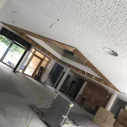 Montaż sufitu podwieszanego z perforowanej płyty w przestronnym biurze, widoczne kable elektryczne i elementy drewniane konstrukcji oraz fragmenty posadzki i mebli zabezpieczonych folią.