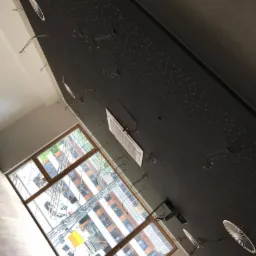 Czarny sufit z perforacją i przygotowanymi otworami na oświetlenie, widoczne wystające kable, kratka wentylacyjna i okno z widokiem na budynek w tle.