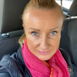 Autoportret kobiety w samochodzie z różowym szalem, widoczny fragment czarnej kurtki i logo na niej.