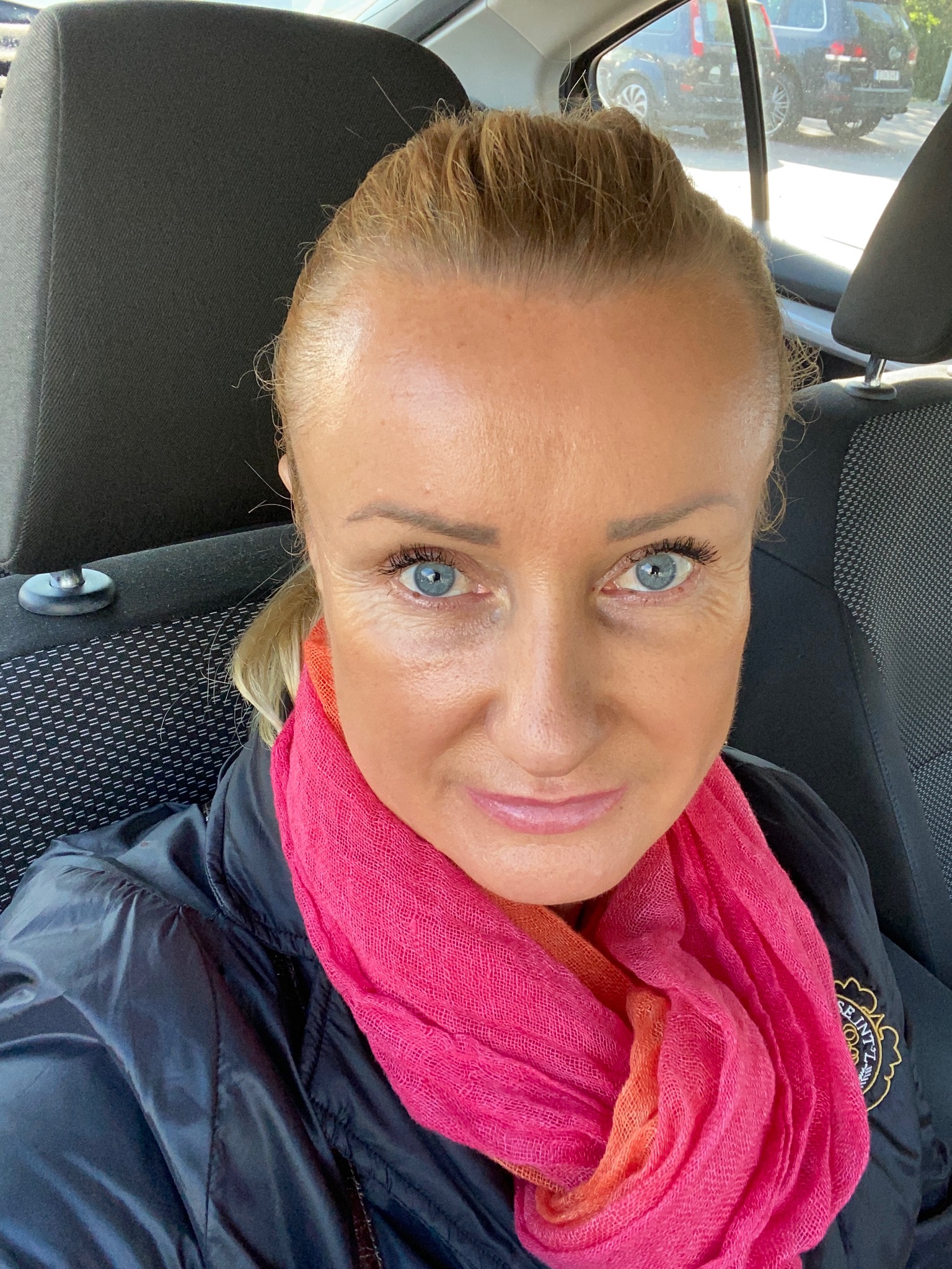 Autoportret kobiety w samochodzie z różowym szalem, widoczny fragment czarnej kurtki i logo na niej.