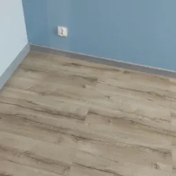 Ułożone panele podłogowe w jasnym odcieniu drewna, widoczne listwy przypodłogowe i fragment ściany pomalowanej na dwa odcienie niebieskiego z gniazdkiem elektrycznym.