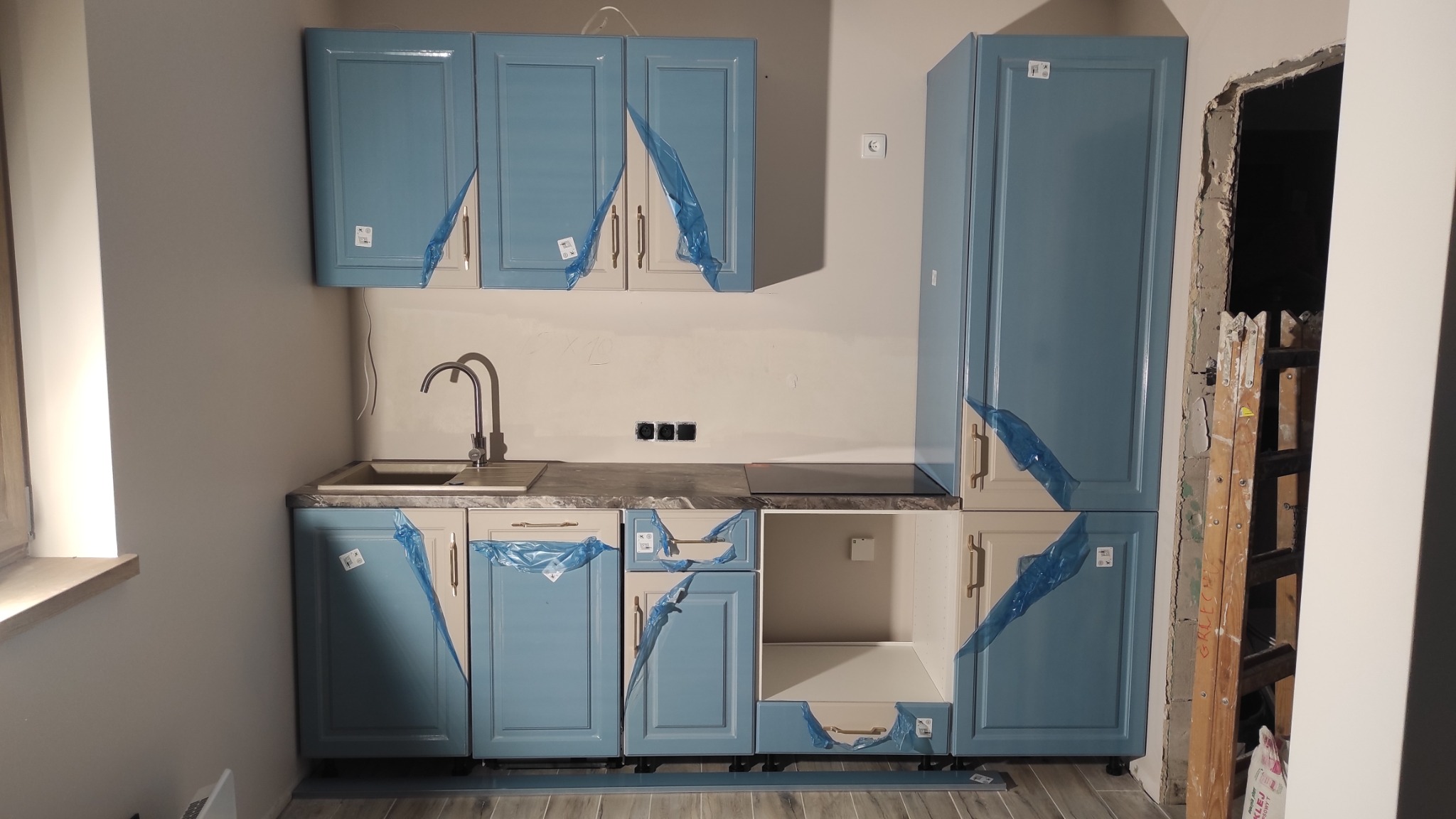 Niebiesko-beżowa zabudowa kuchenna w trakcie montażu, zabezpieczona folią, widoczna drabina oparta o futrynę drzwi.