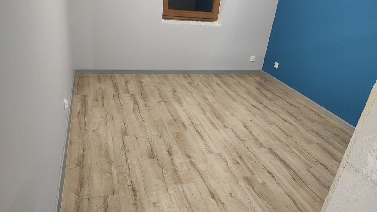 Jasne panele podłogowe ułożone w pokoju w Wejherowie, widoczne fragmenty szarej i niebieskiej ściany, małe okno w drewnianej ramie oraz listwy przypodłogowe.