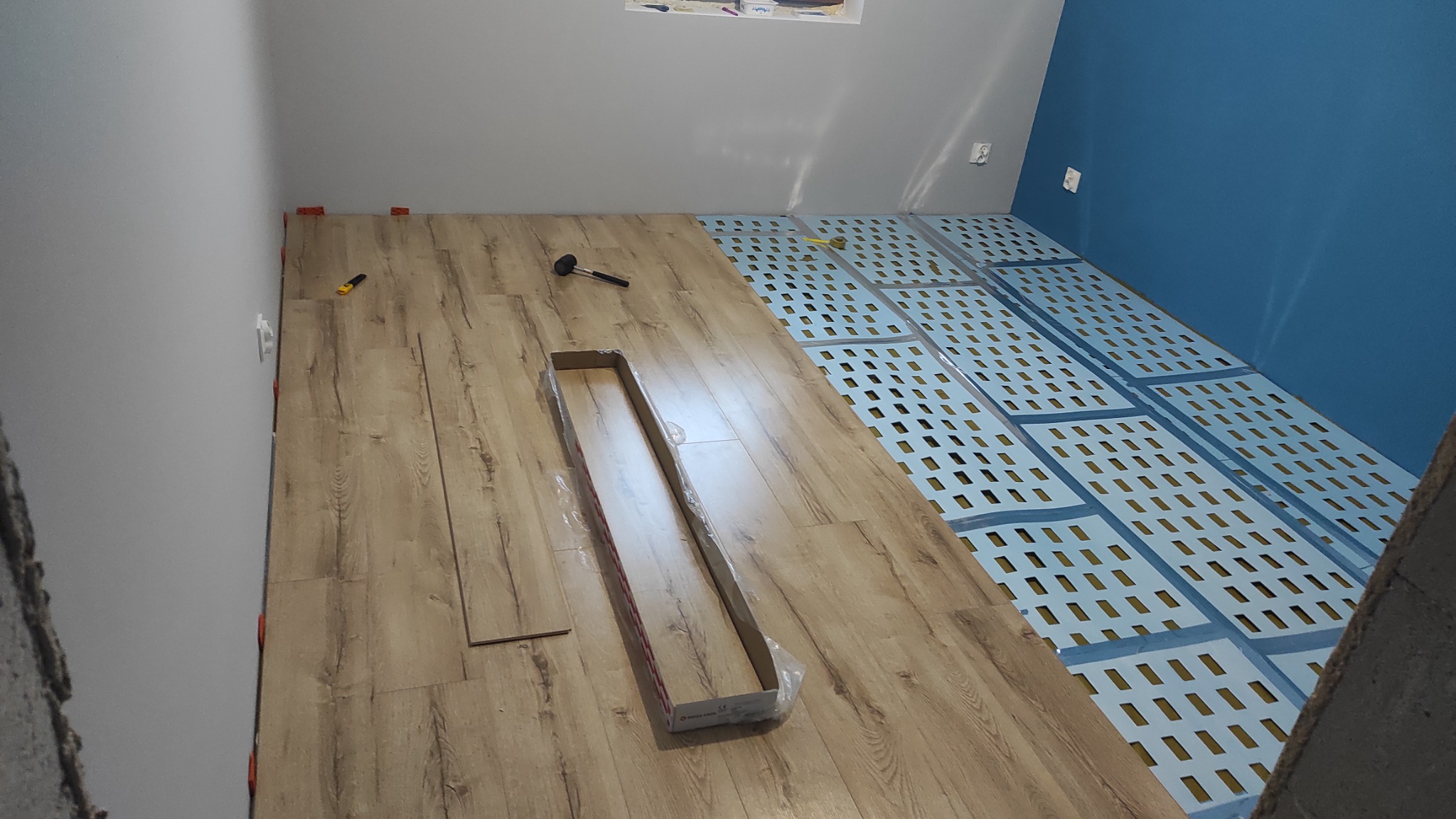 Proces układania paneli podłogowych: widoczne podłoże z folii perforowanej, częściowo ułożone panele imitujące drewno, młotek gumowy i nóż do cięcia, listwa przypodłogowa w opakowaniu.