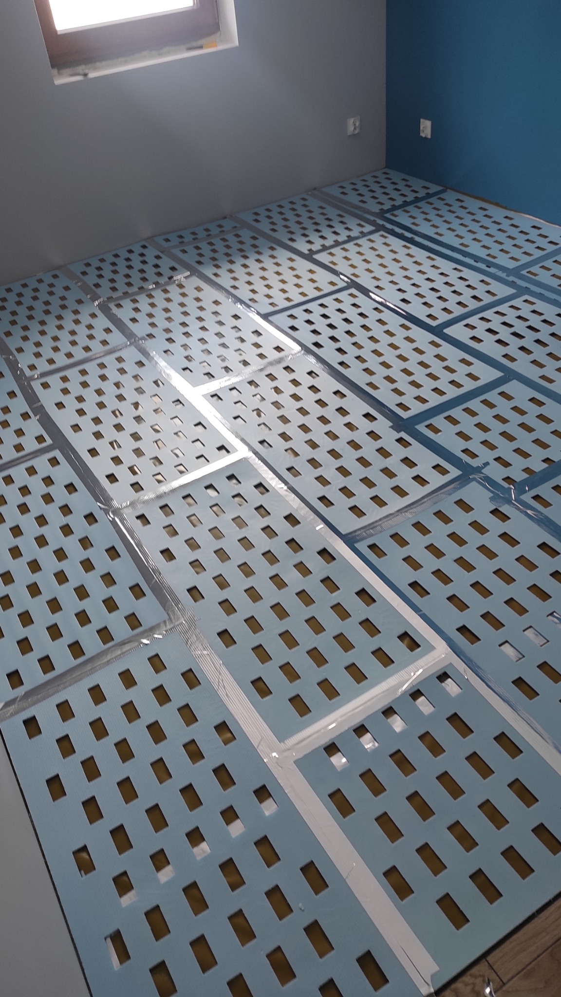 Przygotowane panele podłogowe z otworami, zabezpieczone taśmą, ułożone na podłodze w pokoju z niebieską i szarą ścianą, gotowe do montażu.