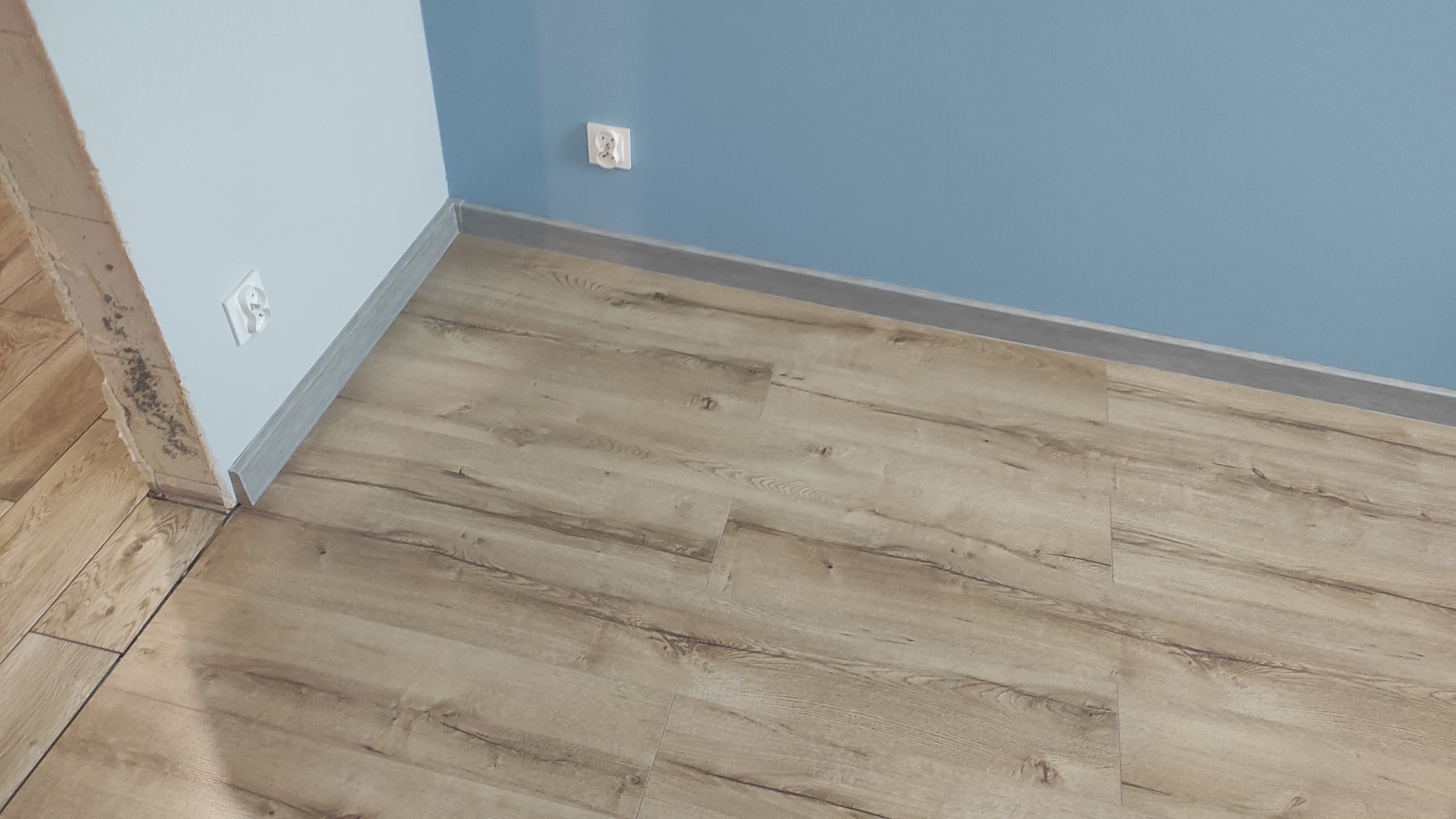 Ułożone panele podłogowe w jasnym odcieniu drewna, widoczne listwy przypodłogowe i fragment ściany pomalowanej na dwa odcienie niebieskiego z gniazdkiem elektrycznym.