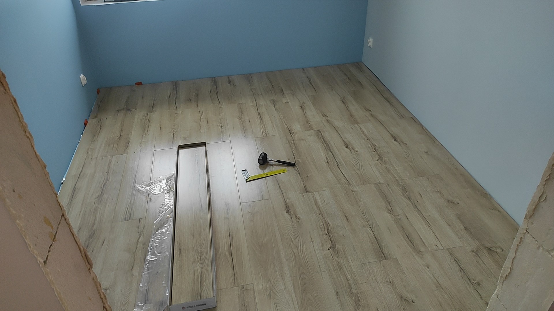 Fragment pokoju z niebieskimi ścianami w trakcie układania podłogi z paneli laminowanych. Widoczne narzędzia: gumowy młotek i kątownik. Część paneli jeszcze w folii.
