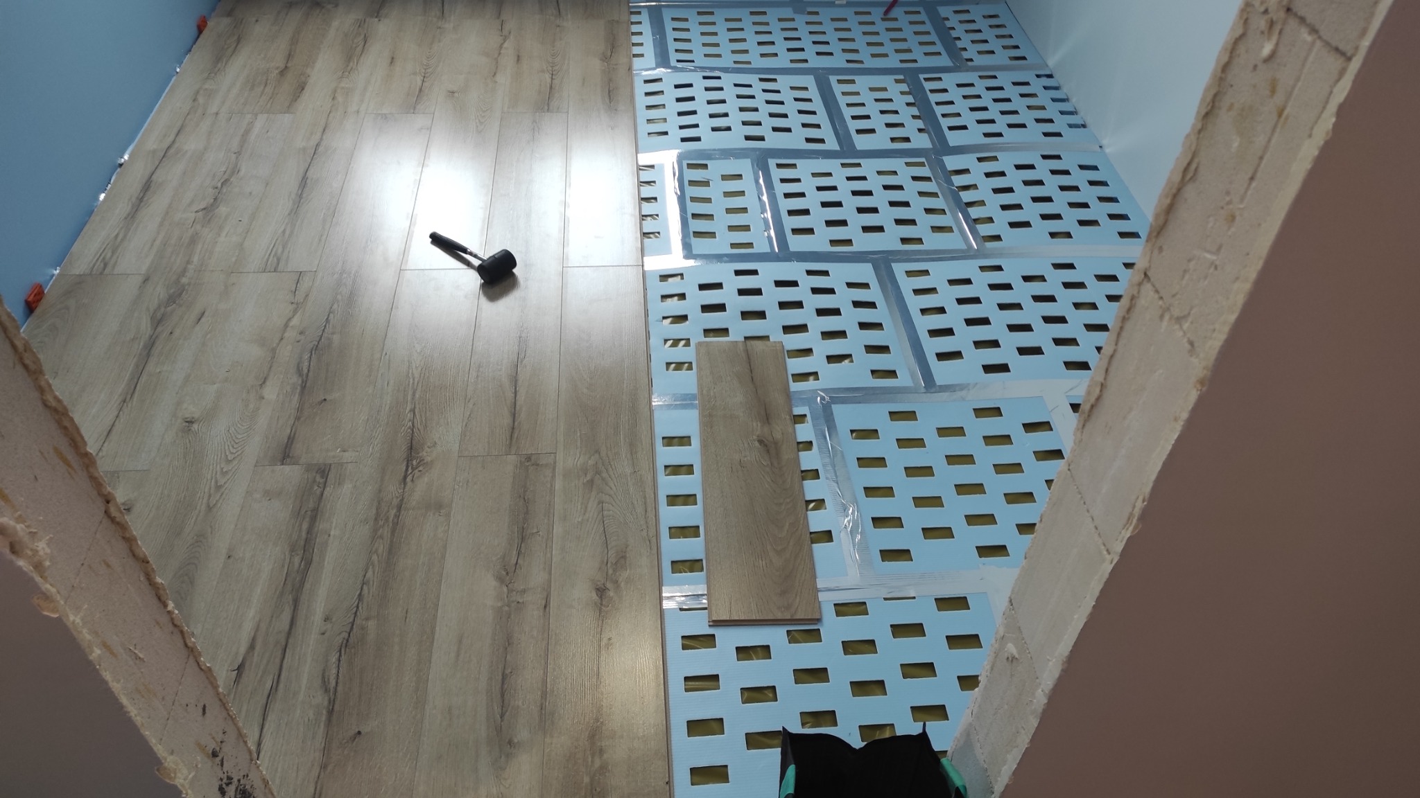 Proces układania paneli podłogowych: widoczna część ułożonej podłogi, mata pod panele z perforacją, pojedynczy panel i gumowy młotek.