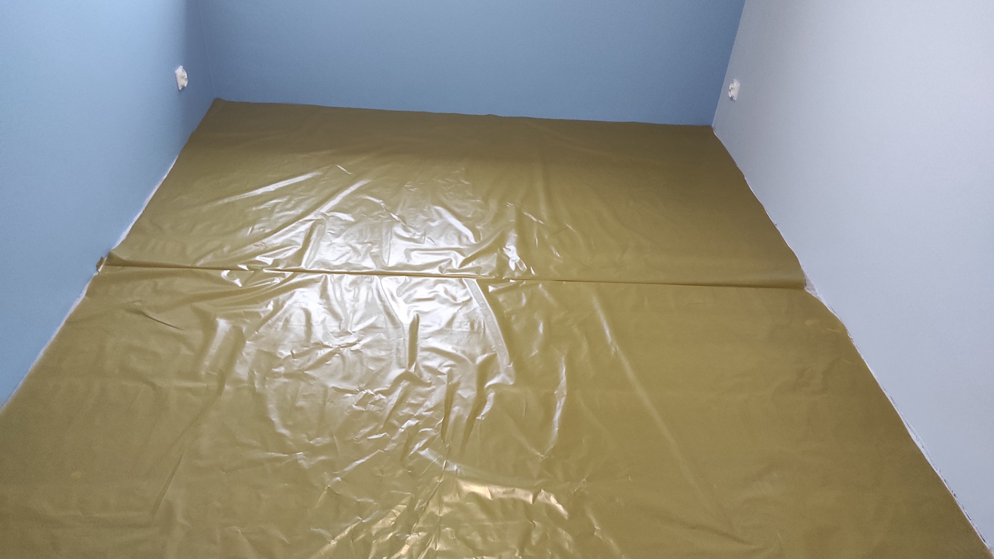 Podłoga w pokoju zabezpieczona folią ochronną przed rozpoczęciem prac remontowych, ściany pomalowane na niebiesko i biało.