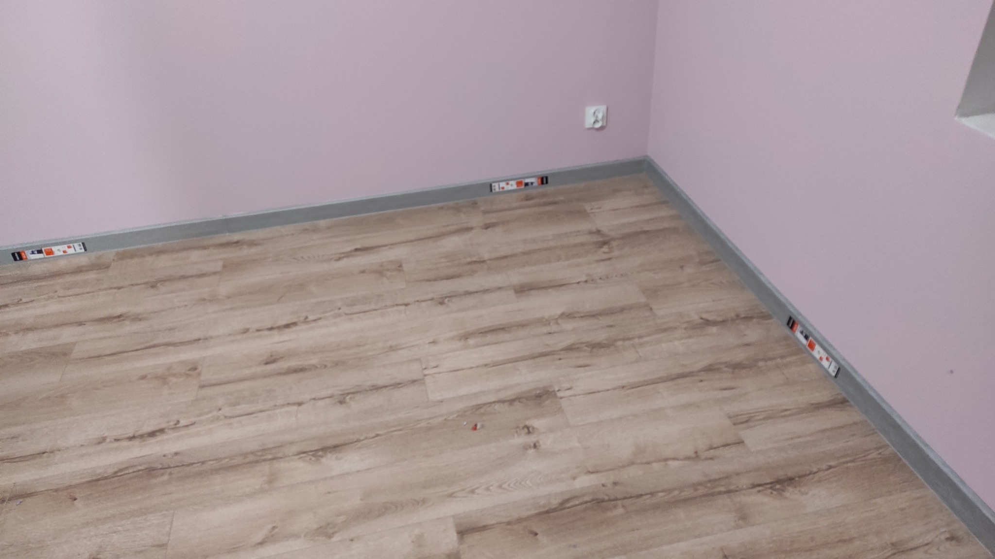 Fragment pokoju z ułożonymi panelami podłogowymi o wzorze drewna, szare listwy przypodłogowe, różowe ściany, widoczne kliny dystansowe przy ścianie.