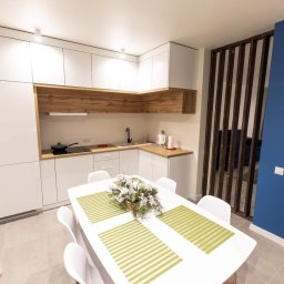 Kristall Sp. z o.o - Jasna kuchnia z białymi szafkami i drewnianymi akcentami, stół z dekoracją kwiatową, niebieski filar z drewnianą ażurową ścianką działową.