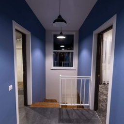 Kristall Sp. z o.o - Korytarz z niebieskimi ścianami, szarymi płytkami podłogowymi i białymi futrynami drzwi, oświetlony wiszącymi lampami i naturalnym światłem z okna.