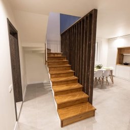 Kristall Sp. z o.o - Drewniane schody z ażurową balustradą w nowoczesnym wnętrzu z widoczną kuchnią i jadalnią w tle, akcent ściany w kolorze granatowym.