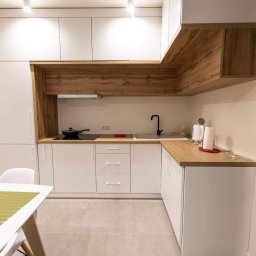 Kristall Sp. z o.o - Nowoczesna kuchnia z białymi szafkami i drewnianymi akcentami, czarny kran i płyta indukcyjna z patelnią, jasne płytki podłogowe, stół z zielonym bieżnikiem.