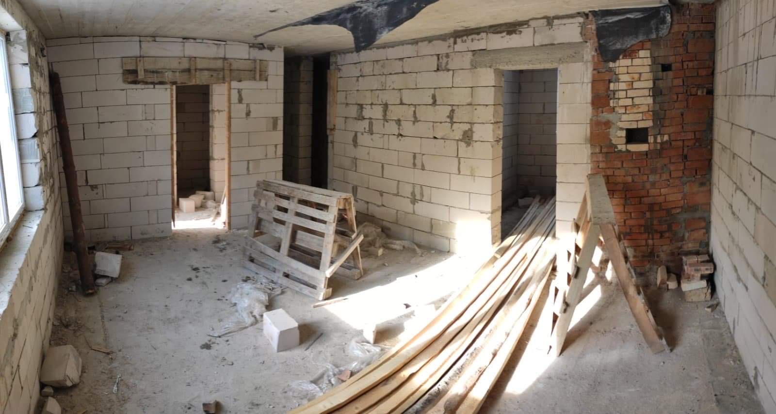 Surowy stan wnętrza w trakcie prac: ściany z białych bloczków, fragment muru z czerwonej cegły, drewniane deski i paleta na podłodze, widoczne otwory drzwiowe.