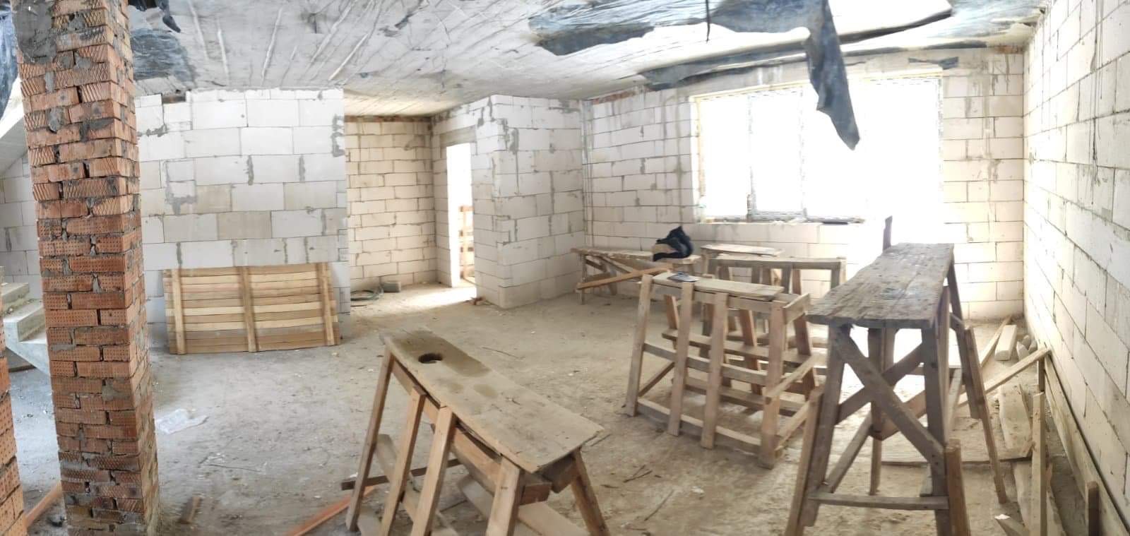 Surowy stan wnętrza w trakcie prac: murowane ściany z bloczków, kolumna z czerwonej cegły, drewniane kozły malarskie i prowizoryczne rusztowania, zabezpieczona folią sufitowa płyta.