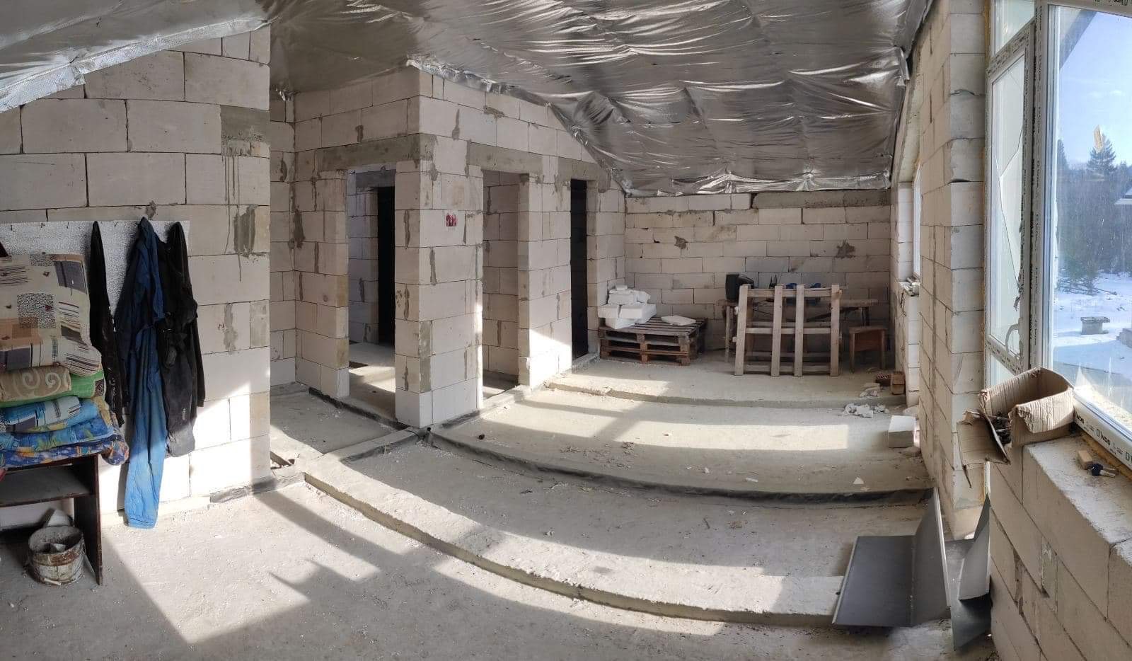 Surowy stan poddasza w trakcie prac wykończeniowych, widoczne mury z białych bloczków, folia izolacyjna na dachu, przygotowane otwory na drzwi i okna, oraz prowizoryczne meble i ubrania.