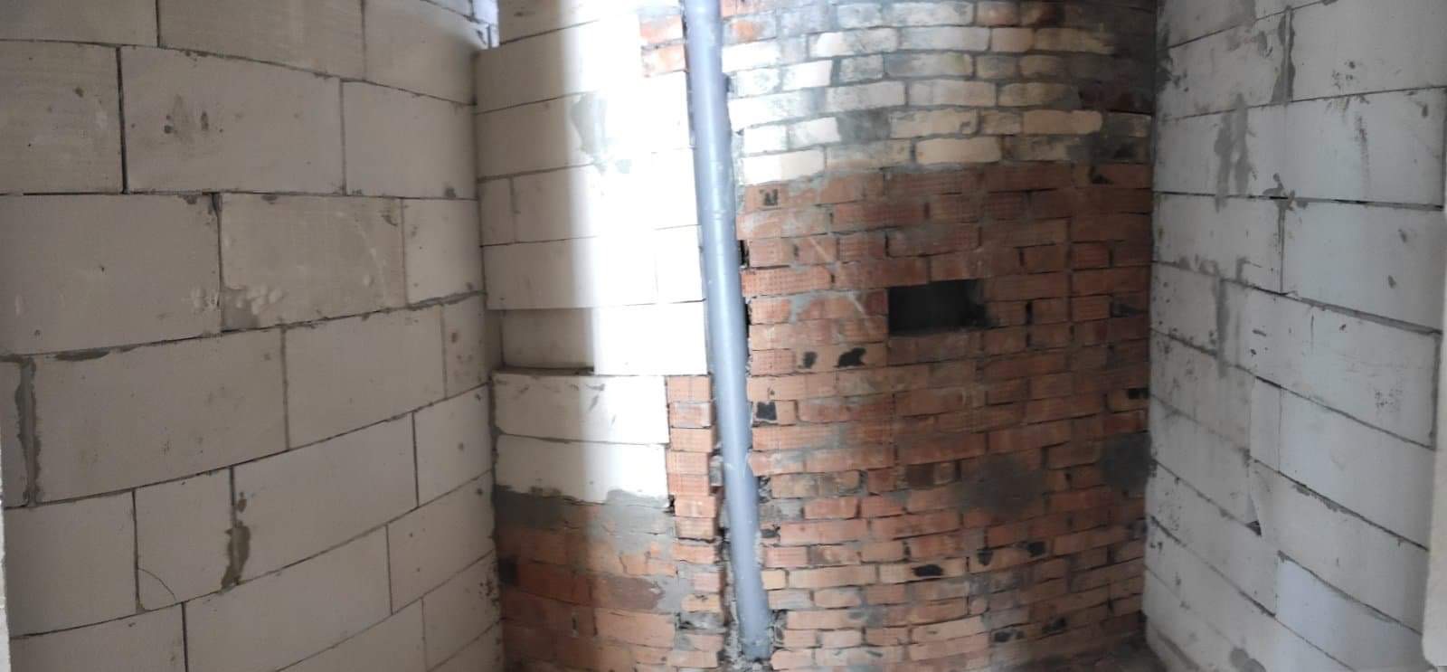 Surowy stan wnętrza: ściany z białych bloczków i fragment ściany z czerwonej cegły z otworem, pionowa rura instalacyjna, etap prac murarskich.