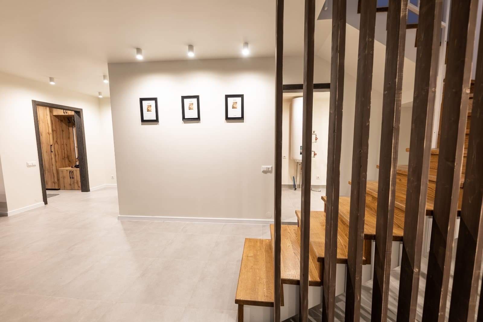 Jasne wnętrze domu z szarymi płytkami podłogowymi, drewnianymi schodami z ażurową balustradą, trzema obrazkami w czarnych ramkach na ścianie i drewnianymi drzwiami z ciemną ościeżnicą.