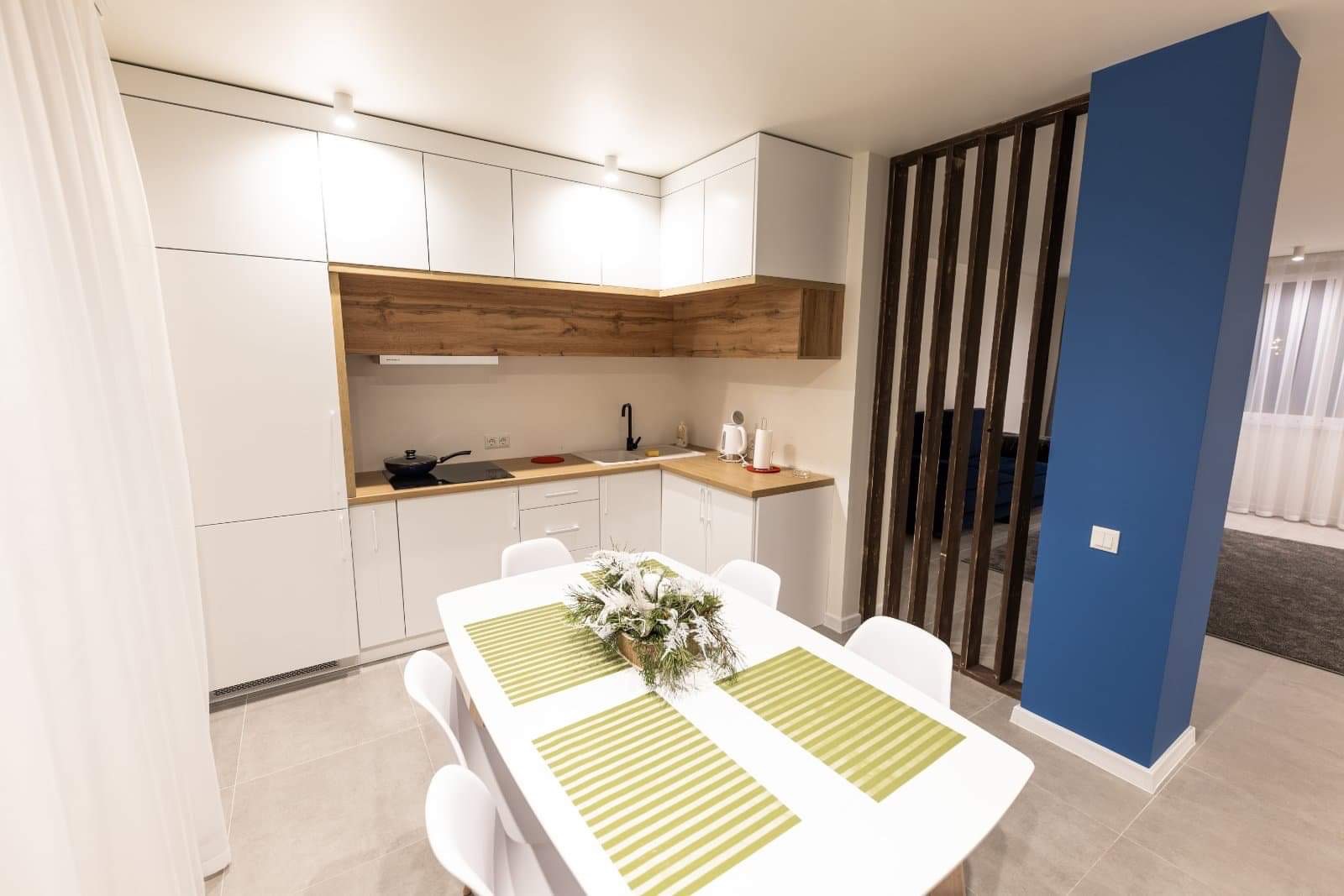 Jasna kuchnia z białymi szafkami i drewnianymi akcentami, stół z dekoracją kwiatową, niebieski filar z drewnianą ażurową ścianką działową.