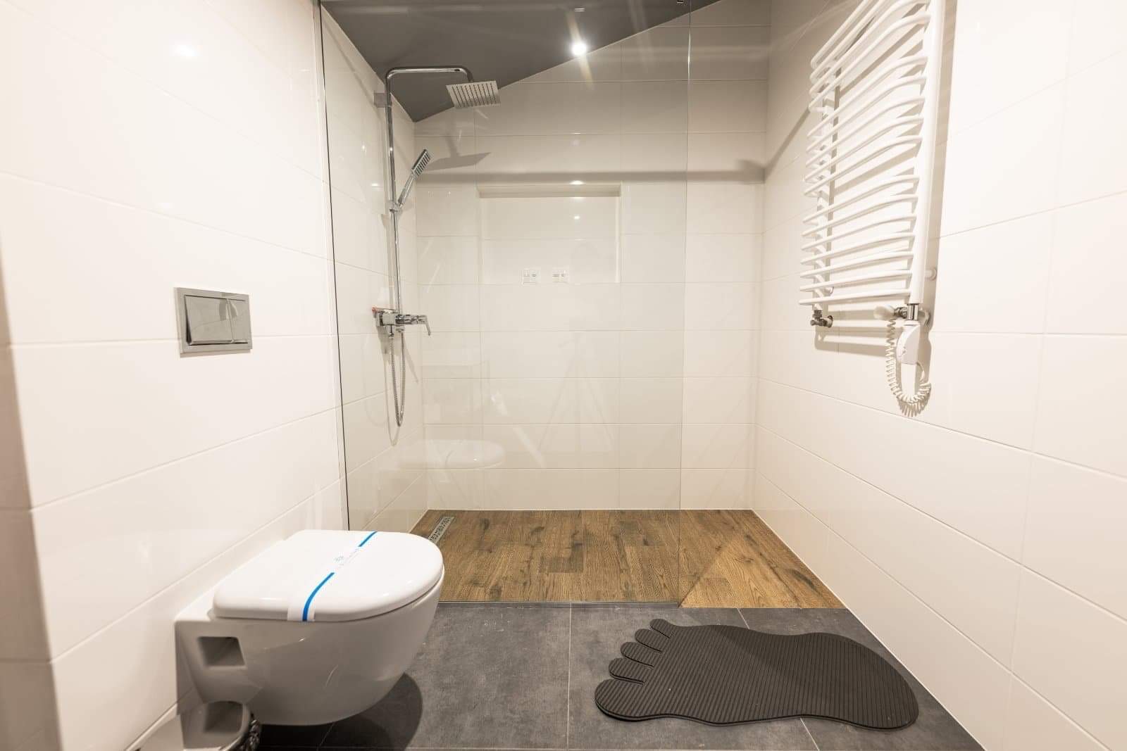 Nowoczesna łazienka z kabiną prysznicową typu walk-in, drewnopodobną podłogą w strefie prysznica, podwieszaną toaletą z niebieskim paskiem higienicznym i grzejnikiem drabinkowym