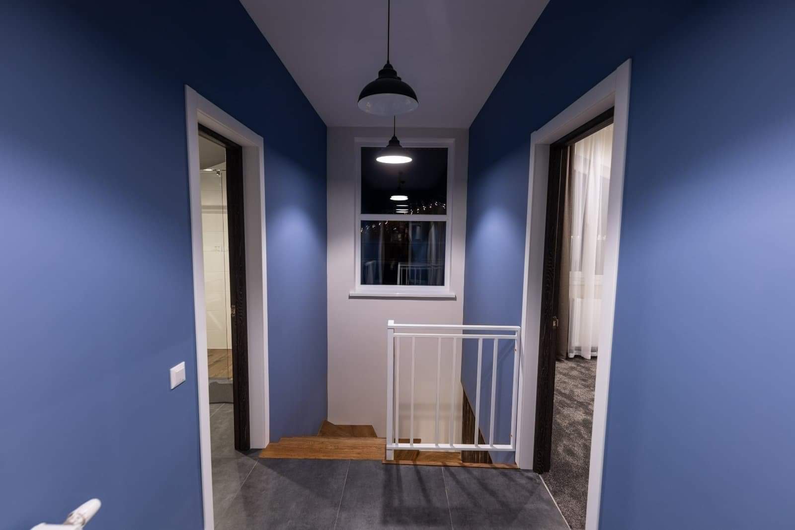 Korytarz z niebieskimi ścianami, szarymi płytkami podłogowymi i białymi futrynami drzwi, oświetlony wiszącymi lampami i naturalnym światłem z okna.