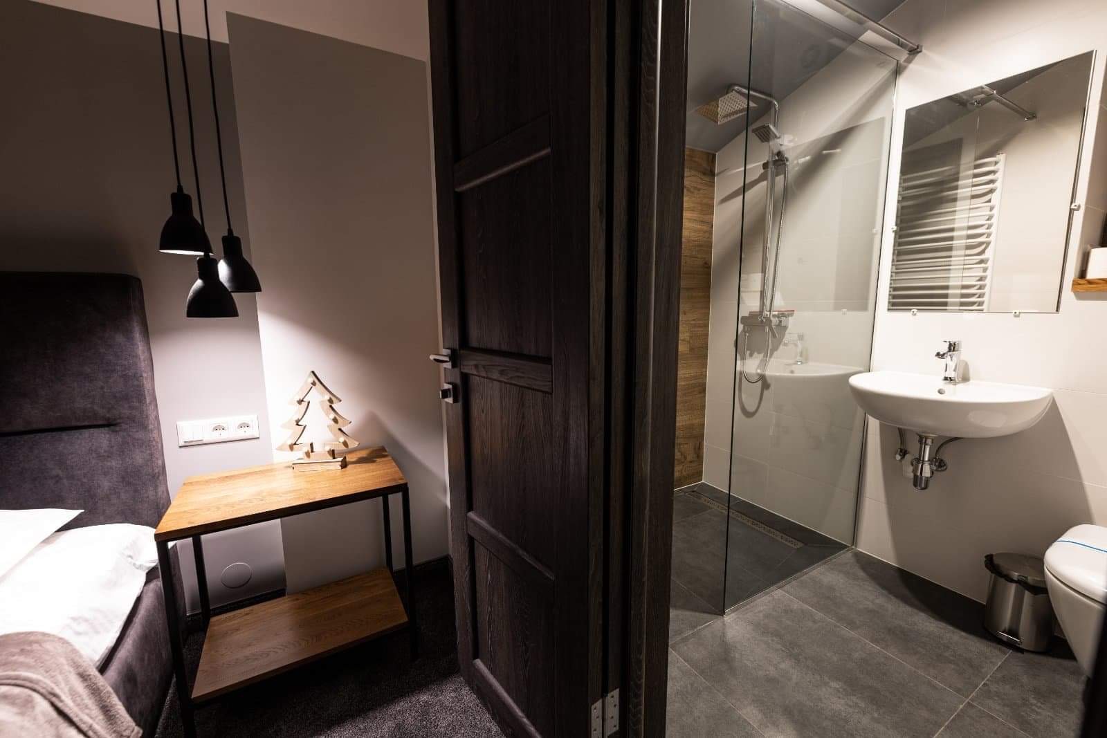 Otwarta, ciemna drewniana stolarka drzwiowa prowadzi do nowoczesnej łazienki z kabiną prysznicową typu walk-in, umywalką nablatową i szarą podłogą; w tle widoczny fragment sypialni z lampami...