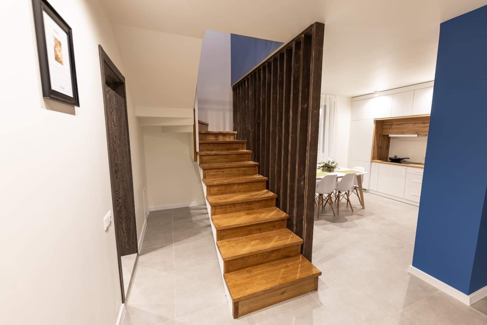 Drewniane schody z ażurową balustradą w nowoczesnym wnętrzu z widoczną kuchnią i jadalnią w tle, akcent ściany w kolorze granatowym.
