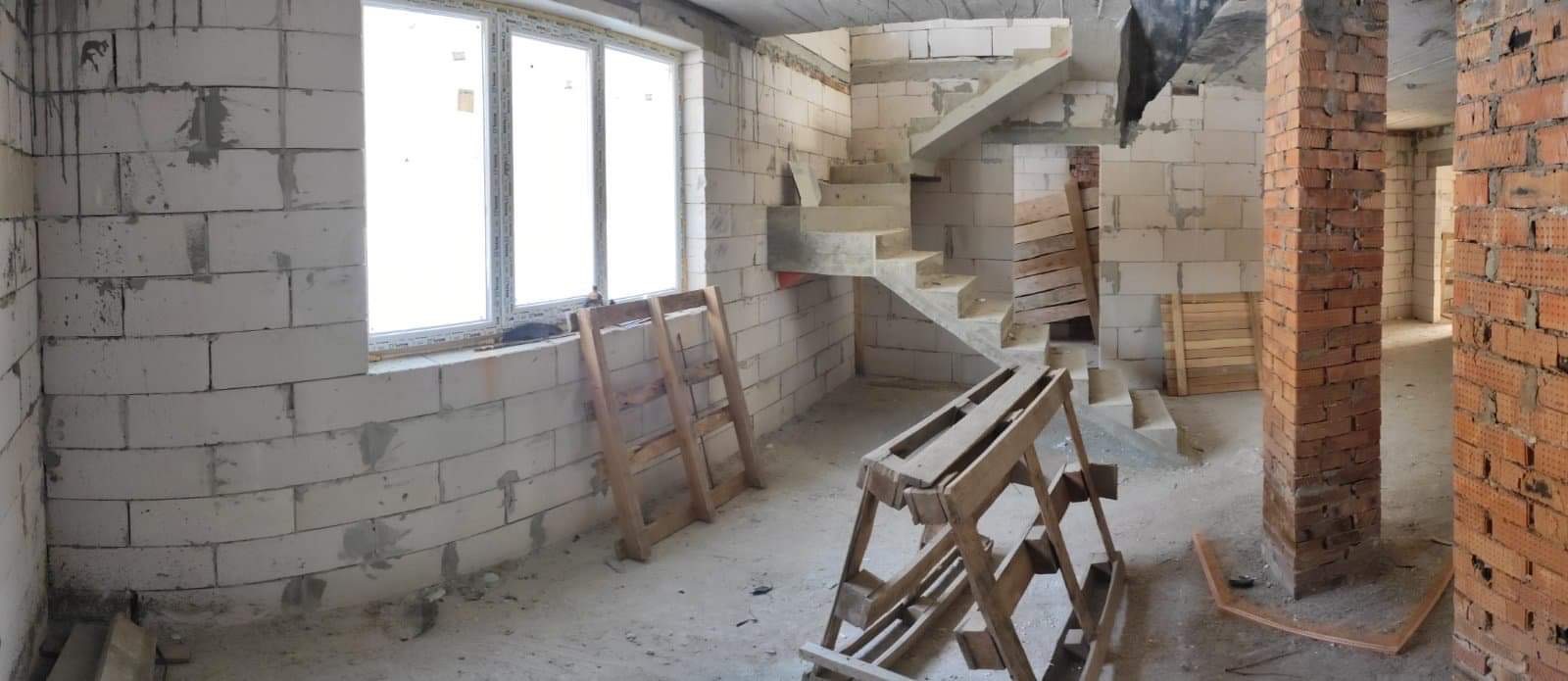 Surowy stan wnętrza w trakcie prac: ściany z białych bloczków, surowa betonowa klatka schodowa, drewniane podpory i rusztowania, kolumna z czerwonej cegły.