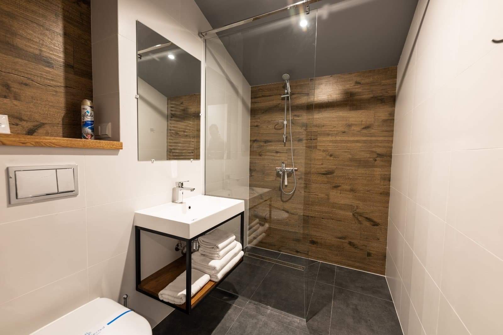 Nowoczesna łazienka z drewnopodobnymi płytkami w kabinie prysznicowej, białą umywalką na czarnym stelażu z drewnianą półką na ręczniki, lustrem i szarą podłogą.