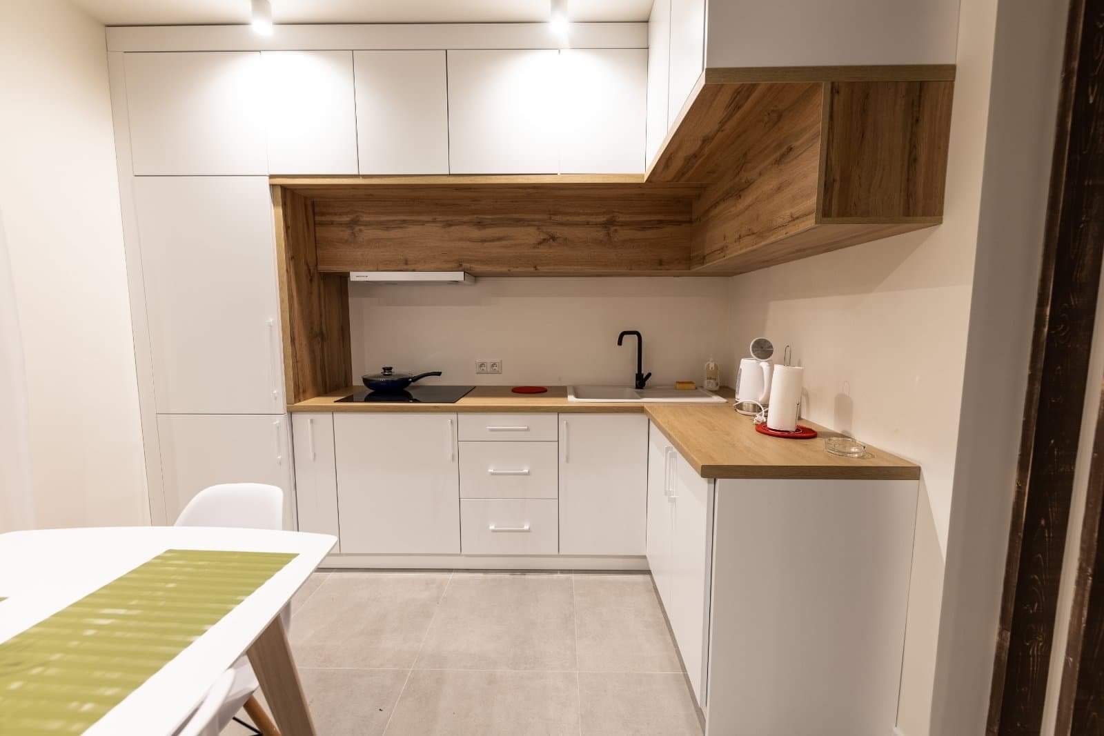 Nowoczesna kuchnia z białymi szafkami i drewnianymi akcentami, czarny kran i płyta indukcyjna z patelnią, jasne płytki podłogowe, stół z zielonym bieżnikiem.