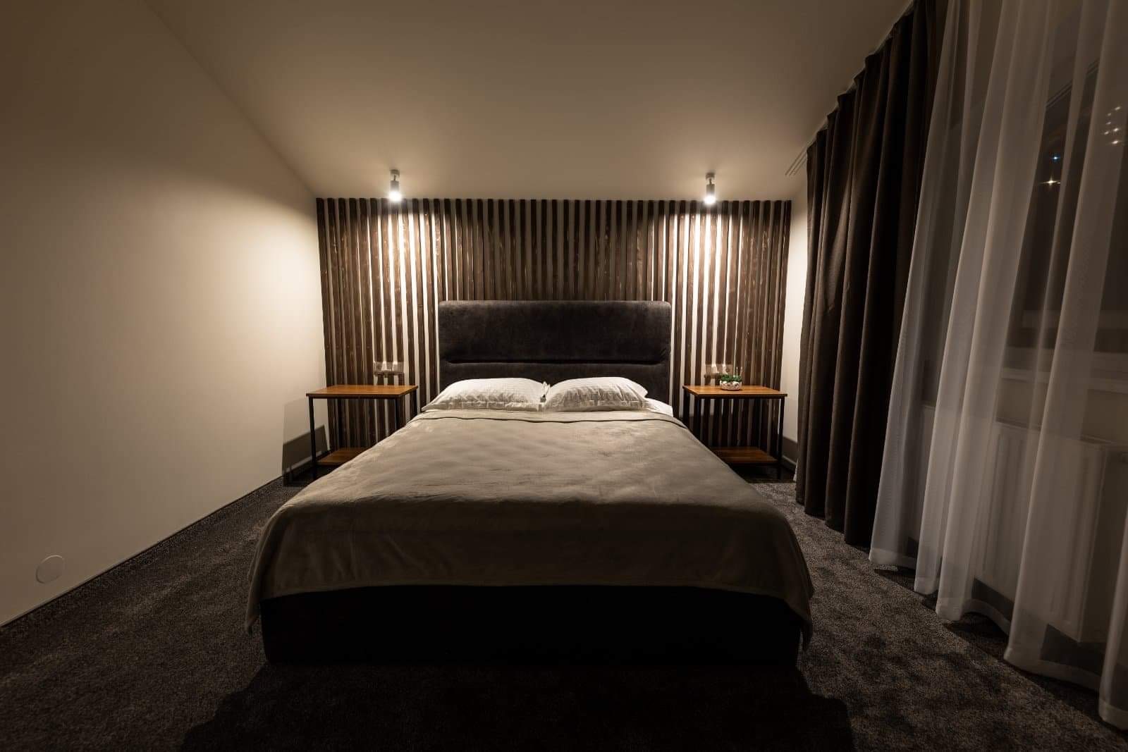 Nowoczesna sypialnia z łóżkiem z szarym zagłówkiem, drewnianą ścianą z lameli, stolikami nocnymi z lampkami i zasłonami w odcieniach brązu i bieli.