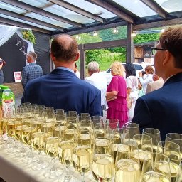 Zagroda Jutrzenki5- winnica, kuchnia, pikniki - Przygotowany bar z rzędami kieliszków wypełnionych szampanem, sokami i wodą, w tle goście weselni i przemawiająca kobieta na tle czarnej ściany z białymi dekoracjami kwiatowymi.