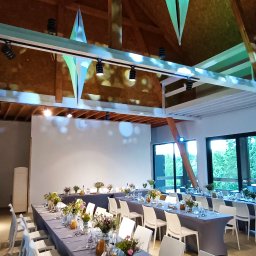 Zagroda Jutrzenki5- winnica, kuchnia, pikniki - Uroczysta sala z długimi stołami przykrytymi szarymi obrusami, nakryta zastawa i kwiatowe dekoracje na stołach, nowoczesne białe krzesła, drewniana konstrukcja dachu z wiszącymi, podświetlonymi...