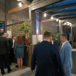Zagroda Jutrzenki5- winnica, kuchnia, pikniki - Uczestnicy w strojach formalnych rozmawiają podczas wydarzenia pod zadaszoną konstrukcją z drewnianymi belkami i dekoracyjnymi lampkami, widoczny bar z menu i kwiatami.