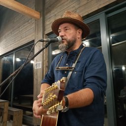 Zagroda Jutrzenki5- winnica, kuchnia, pikniki - Mężczyzna z brodą w kapeluszu śpiewa do mikrofonu i gra na gitarze akustycznej, dodatkowo ma zawieszony na szyi harmonijkę ustną.