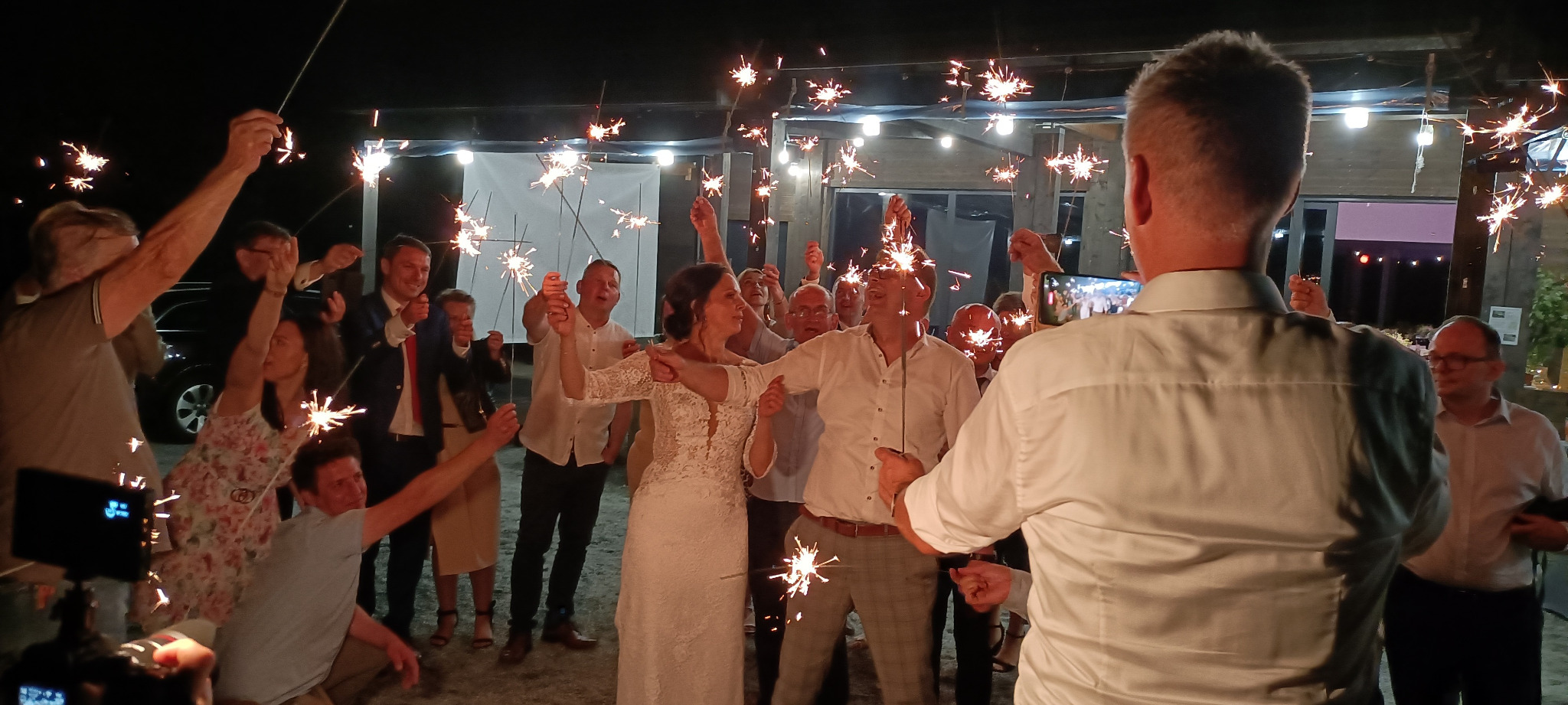 Para młoda otoczona gośćmi weselnymi świętuje z zimnymi ogniami, w tle oświetlony budynek.