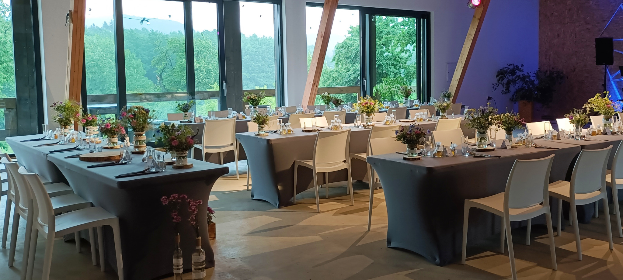 Sala bankietowa z długimi stołami nakrytymi szarymi obrusami, udekorowanymi bukietami polnych kwiatów, szklanymi naczyniami i drewnianymi podkładkami, z widokiem na zielony las za oknami.