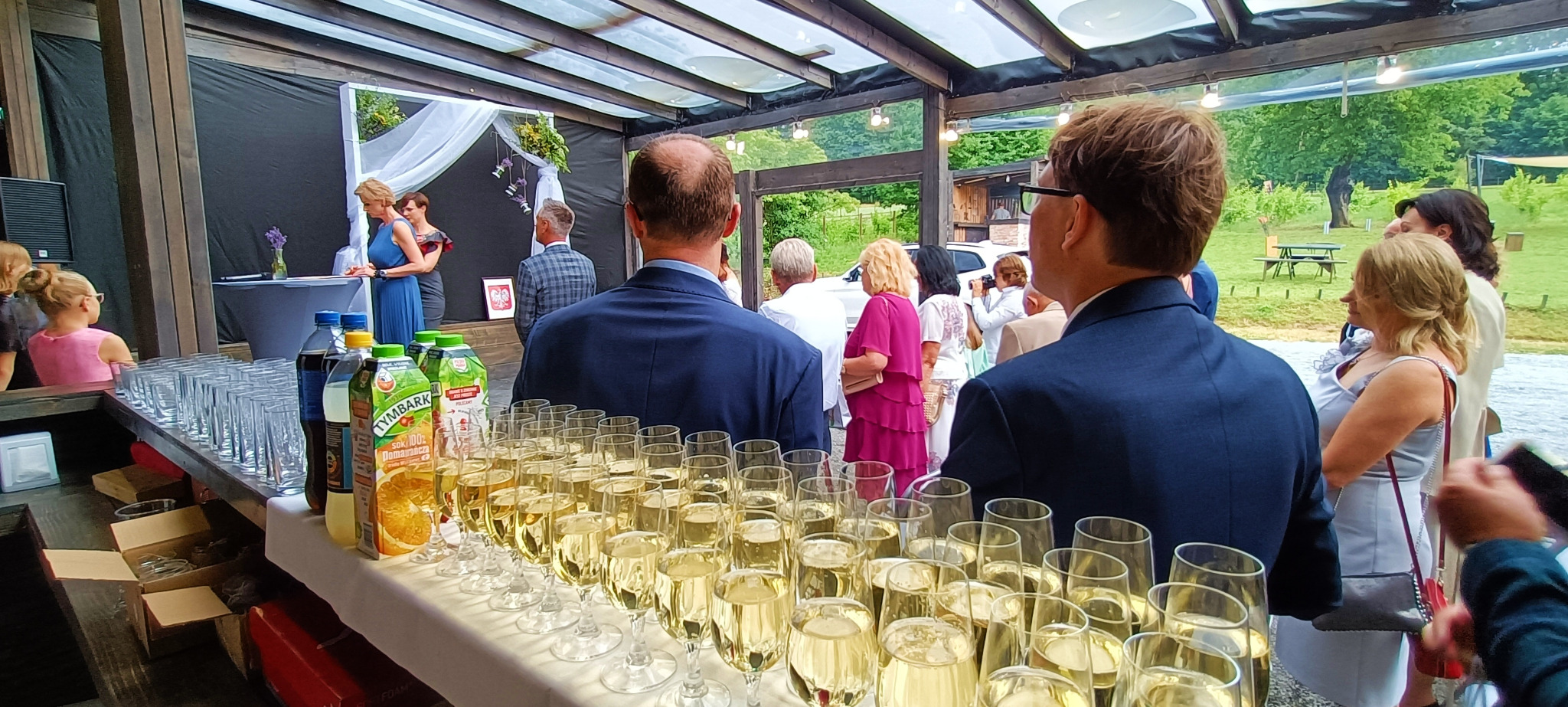 Przygotowany bar z rzędami kieliszków wypełnionych szampanem, sokami i wodą, w tle goście weselni i przemawiająca kobieta na tle czarnej ściany z białymi dekoracjami kwiatowymi.