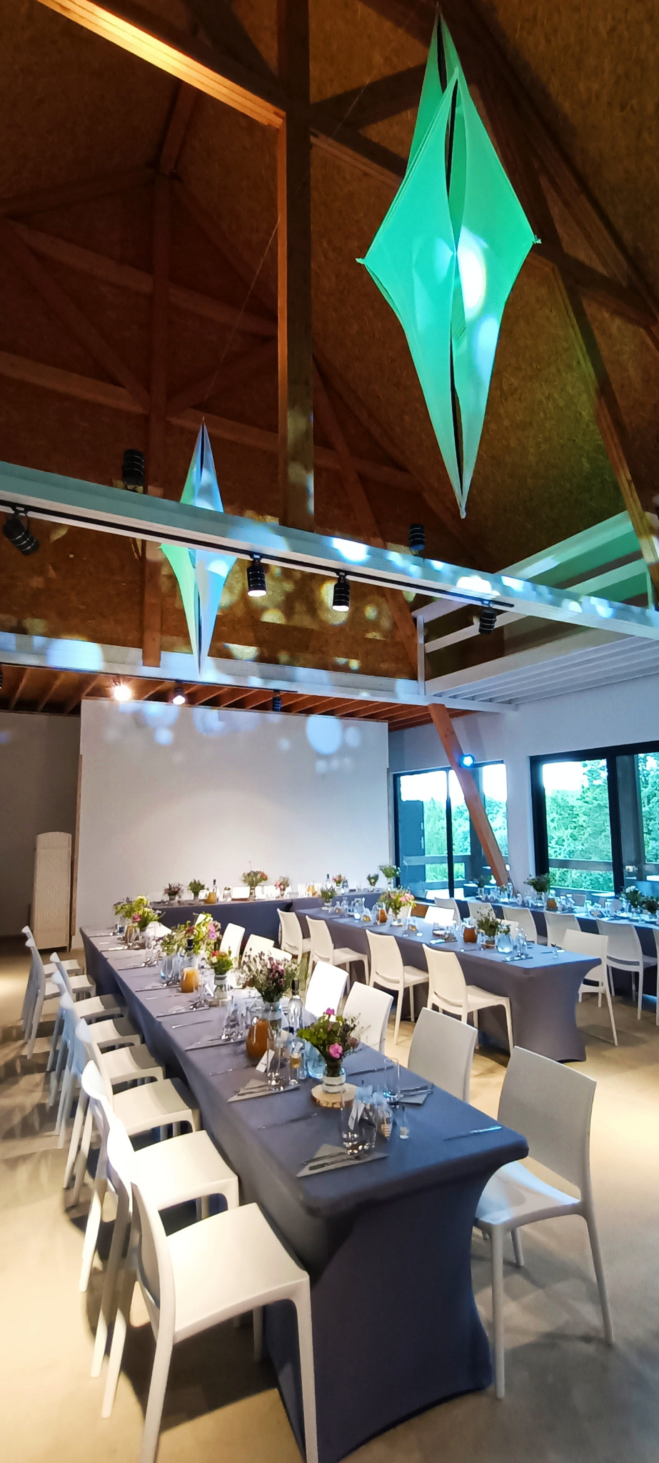 Uroczysta sala z długimi stołami przykrytymi szarymi obrusami, nakryta zastawa i kwiatowe dekoracje na stołach, nowoczesne białe krzesła, drewniana konstrukcja dachu z wiszącymi, podświetlonymi...