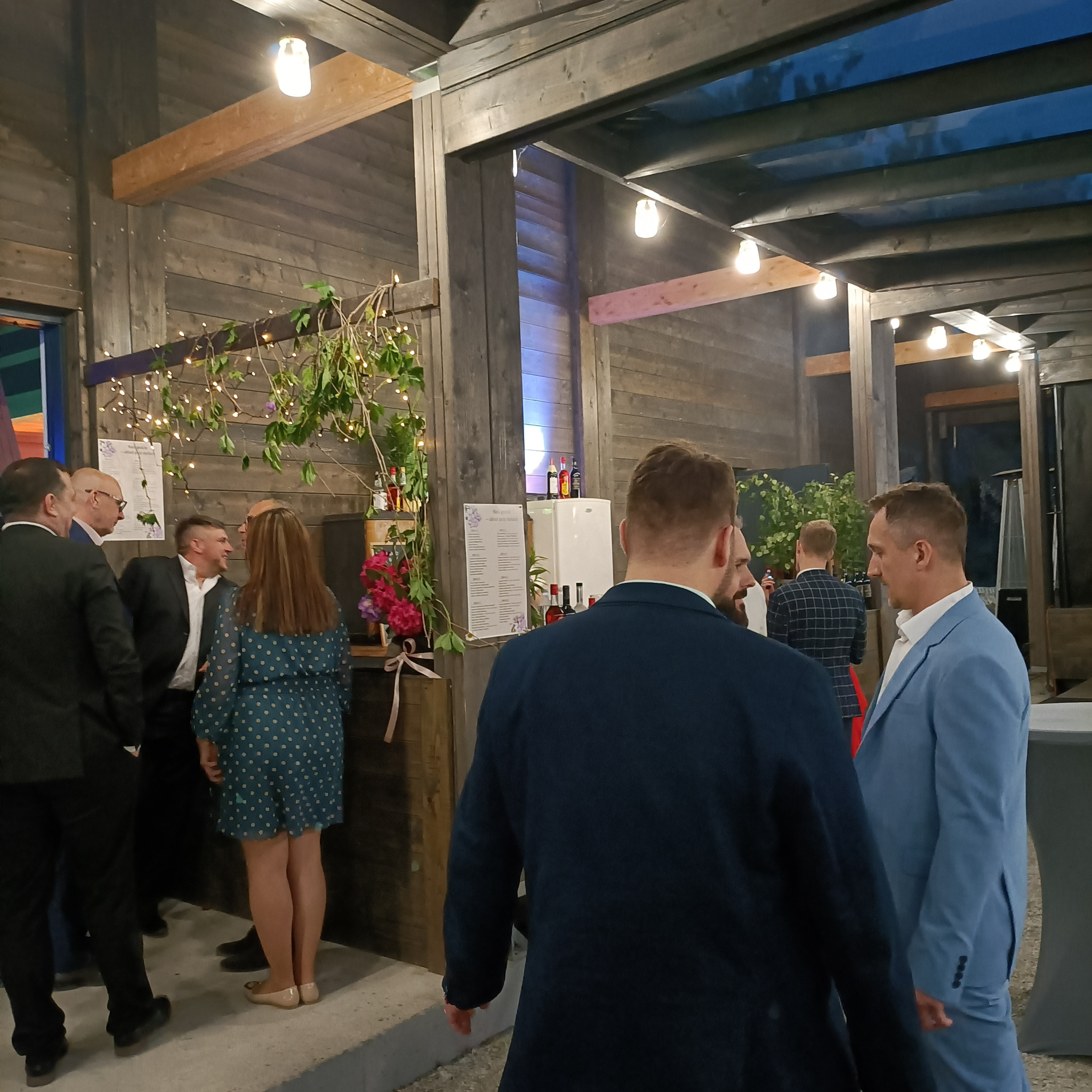 Uczestnicy w strojach formalnych rozmawiają podczas wydarzenia pod zadaszoną konstrukcją z drewnianymi belkami i dekoracyjnymi lampkami, widoczny bar z menu i kwiatami.