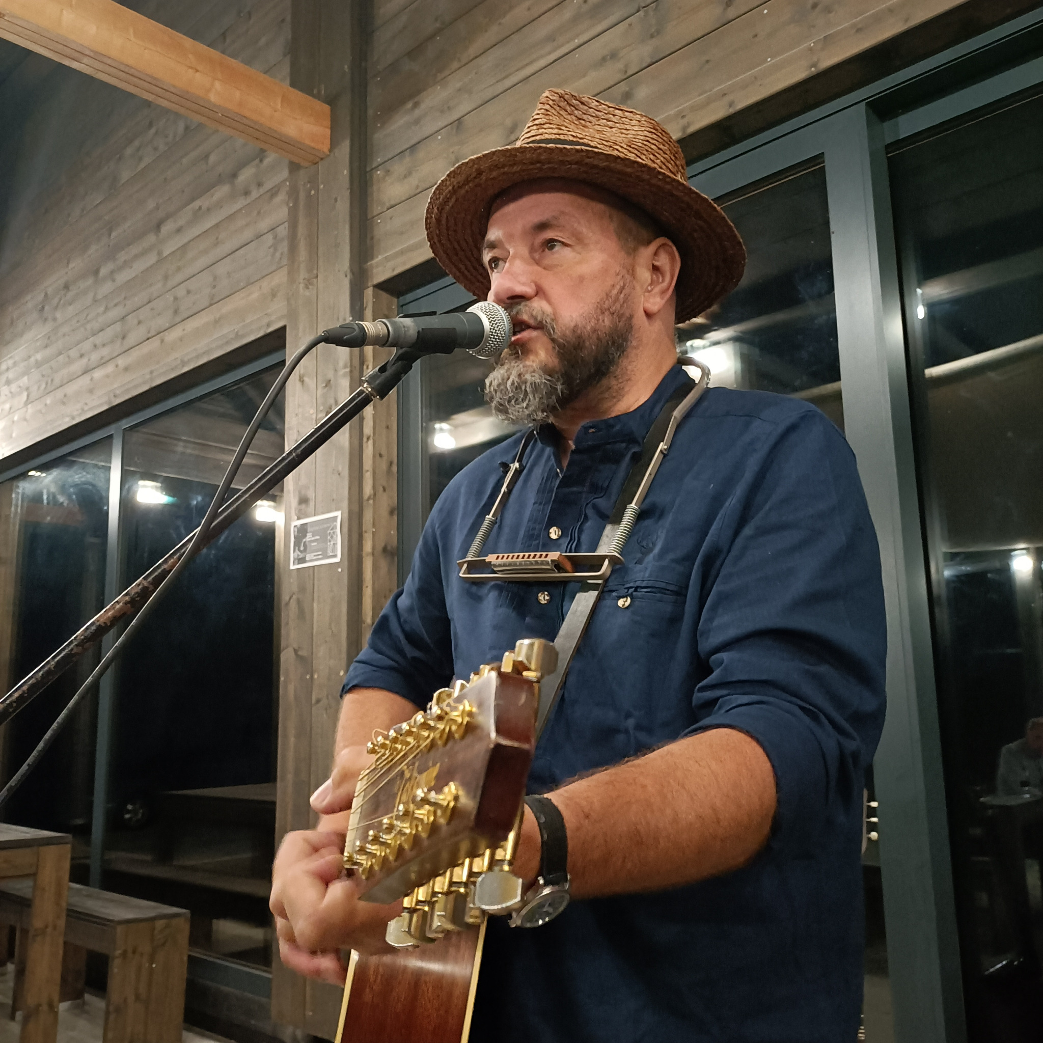 Mężczyzna z brodą w kapeluszu śpiewa do mikrofonu i gra na gitarze akustycznej, dodatkowo ma zawieszony na szyi harmonijkę ustną.